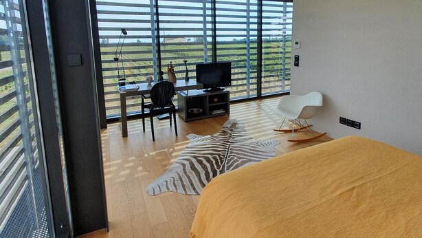 Un dormitorio moderno con un ventanal grande, una cama, un escritorio con una silla y un televisor.
