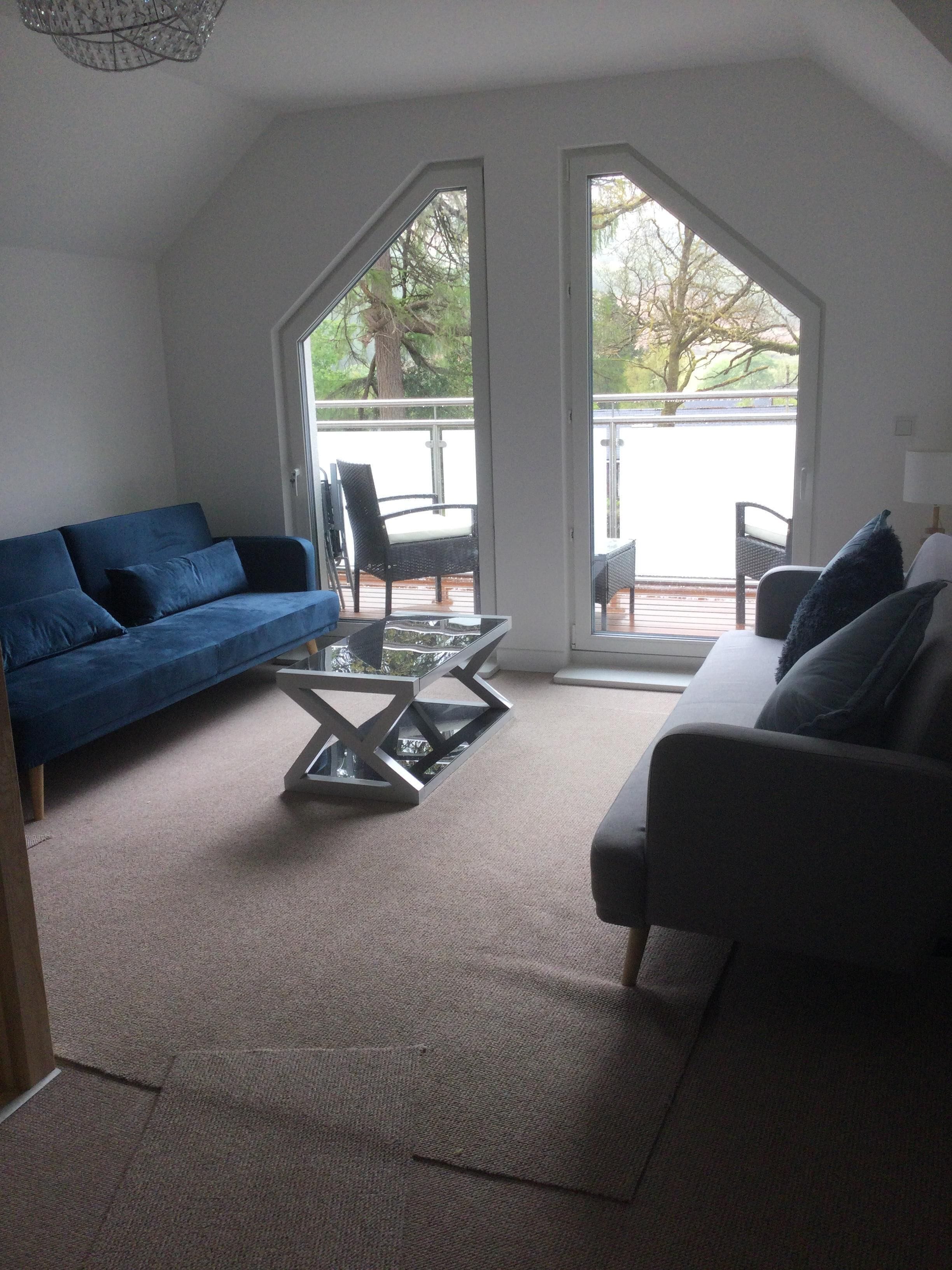 A modern living room with a blue sofa, a grey sofa, and a glass coffee table. There are two large windows providing a view of the outdoors.