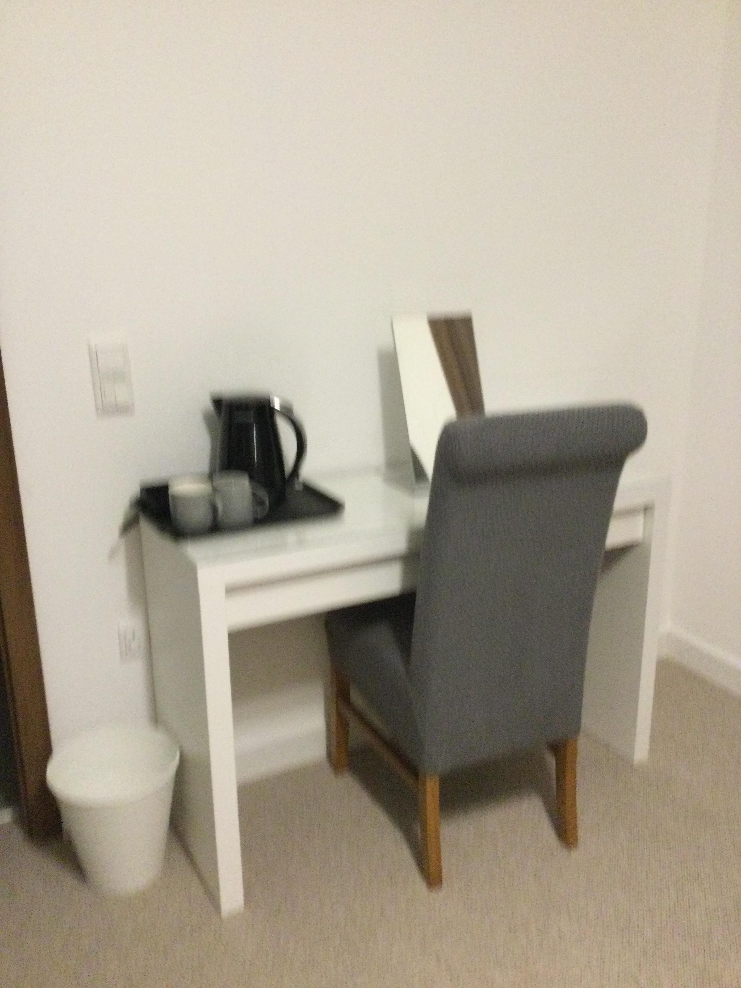 A small, white desk with a grey chair, a black coffee maker, and a white trash bin.