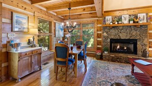 L’intérieur chaleureux d’un chalet avec une cheminée en pierre, une table à manger en bois et une grande fenêtre donnant sur un paysage verdoyant.