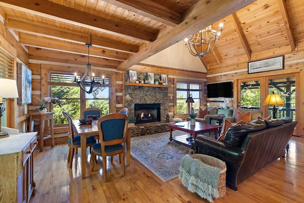 A cozy cabin interior with a stone fireplace, wooden furniture, and large windows offering a view of the outdoors.