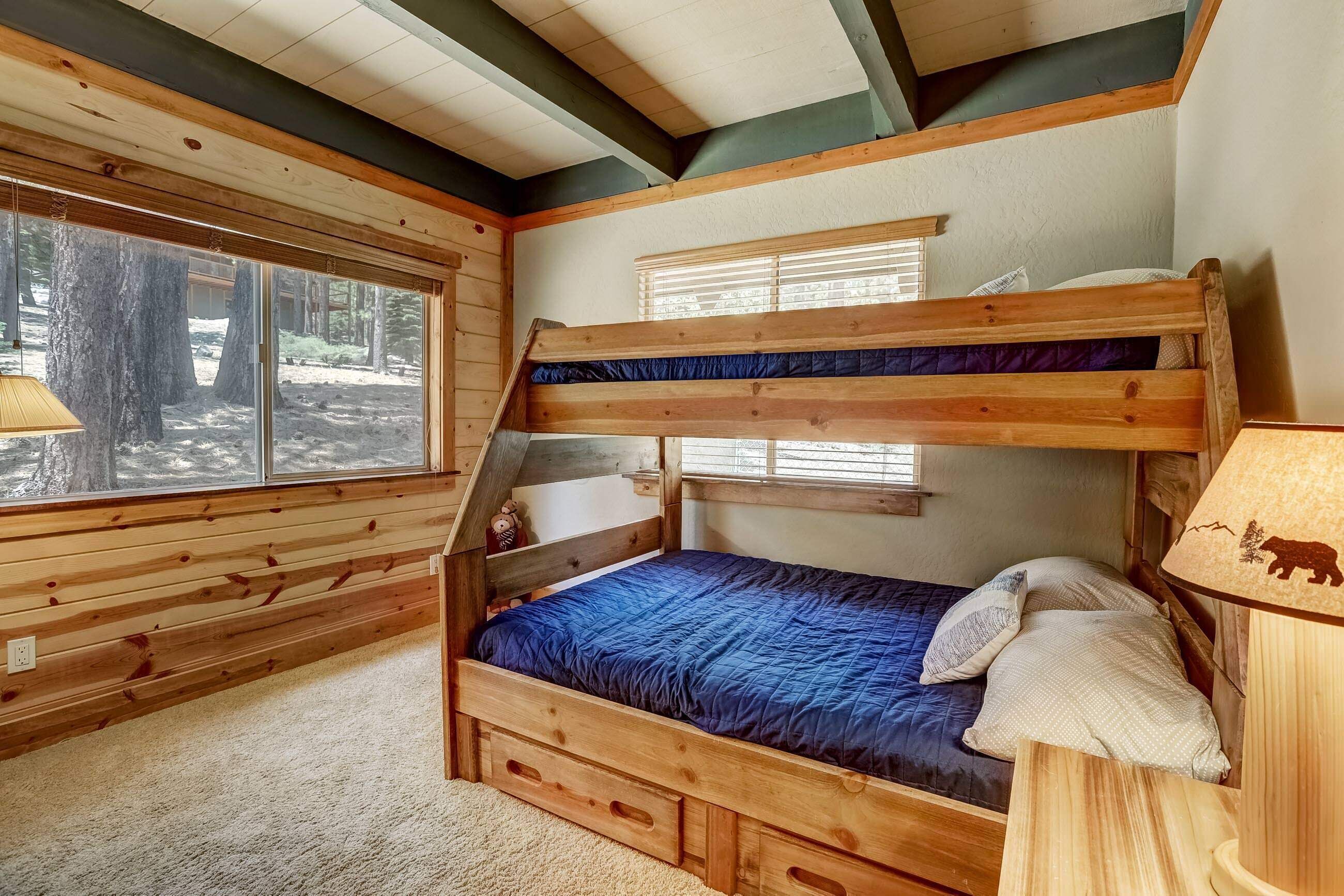Een stapelbed met een blauwe matras en kussens, houten wanden, een raam met zonwering en een lamp met een berendesign.