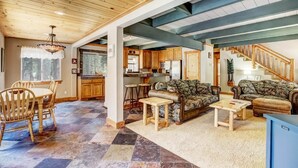 Amplia sala de estar con techo de madera, comedor con mesa y sillas, y cocina con muebles de madera y una escalera.