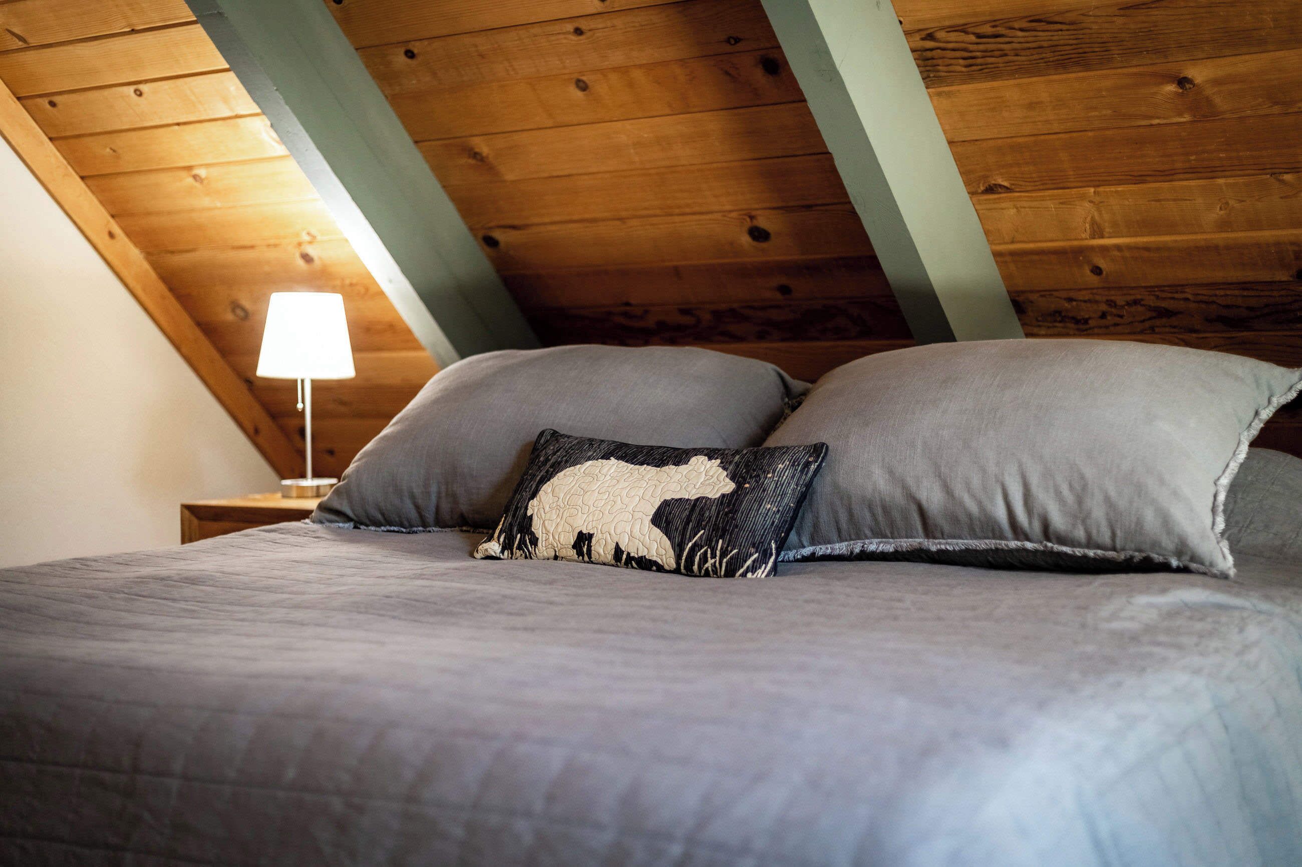 A bedroom with a wooden ceiling, a bed with grey bedding, and a lamp on a nightstand.