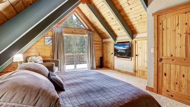 Un dormitorio con techo inclinado, una cama con cubrecama acolchado, un televisor en la pared y una puerta de madera.