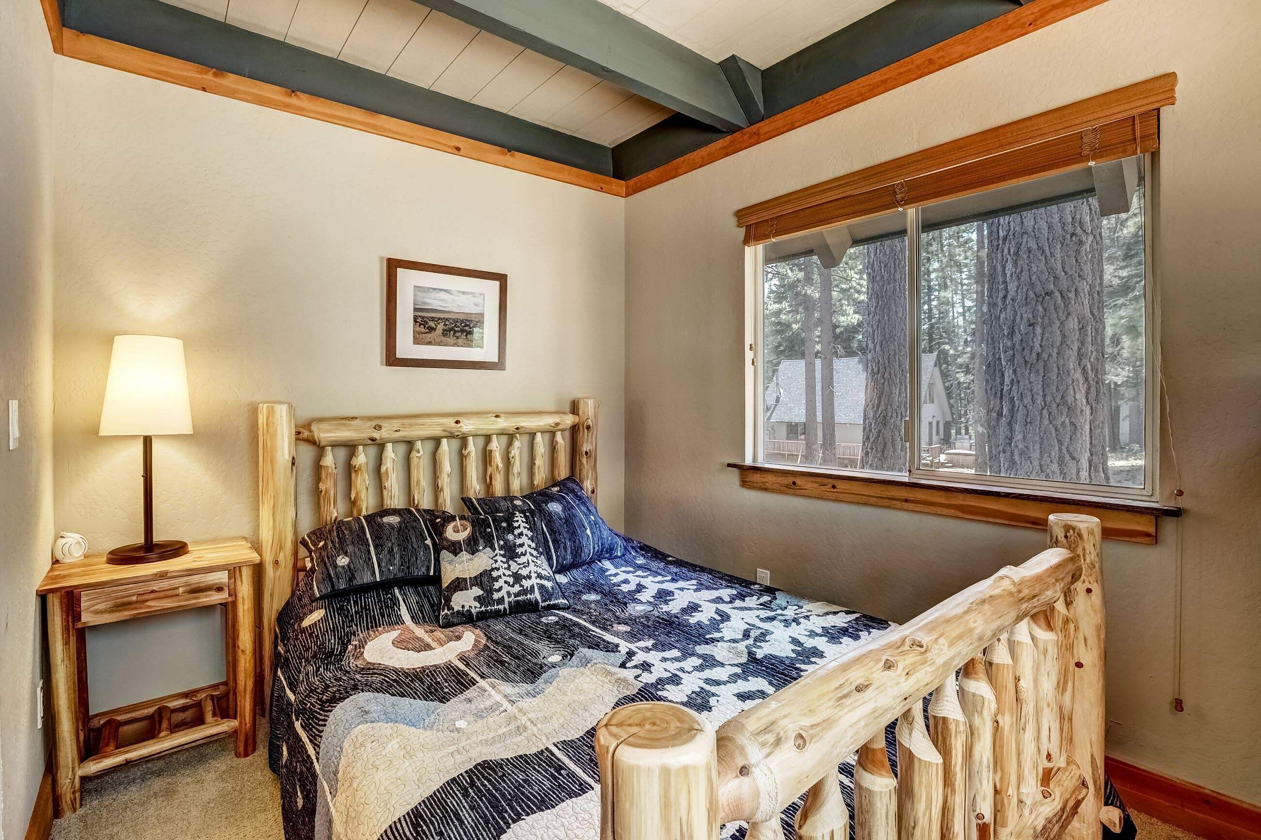 A bedroom with a wooden bed, a nightstand with a lamp, a framed picture on the wall, and a window with a view of trees.