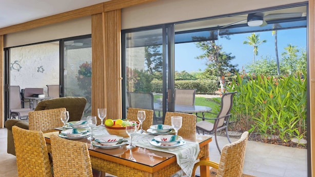 Un comedor con una mesa dispuesta para seis personas, rodeado de sillas de mimbre, y vistas al exterior con follaje y palmeras.