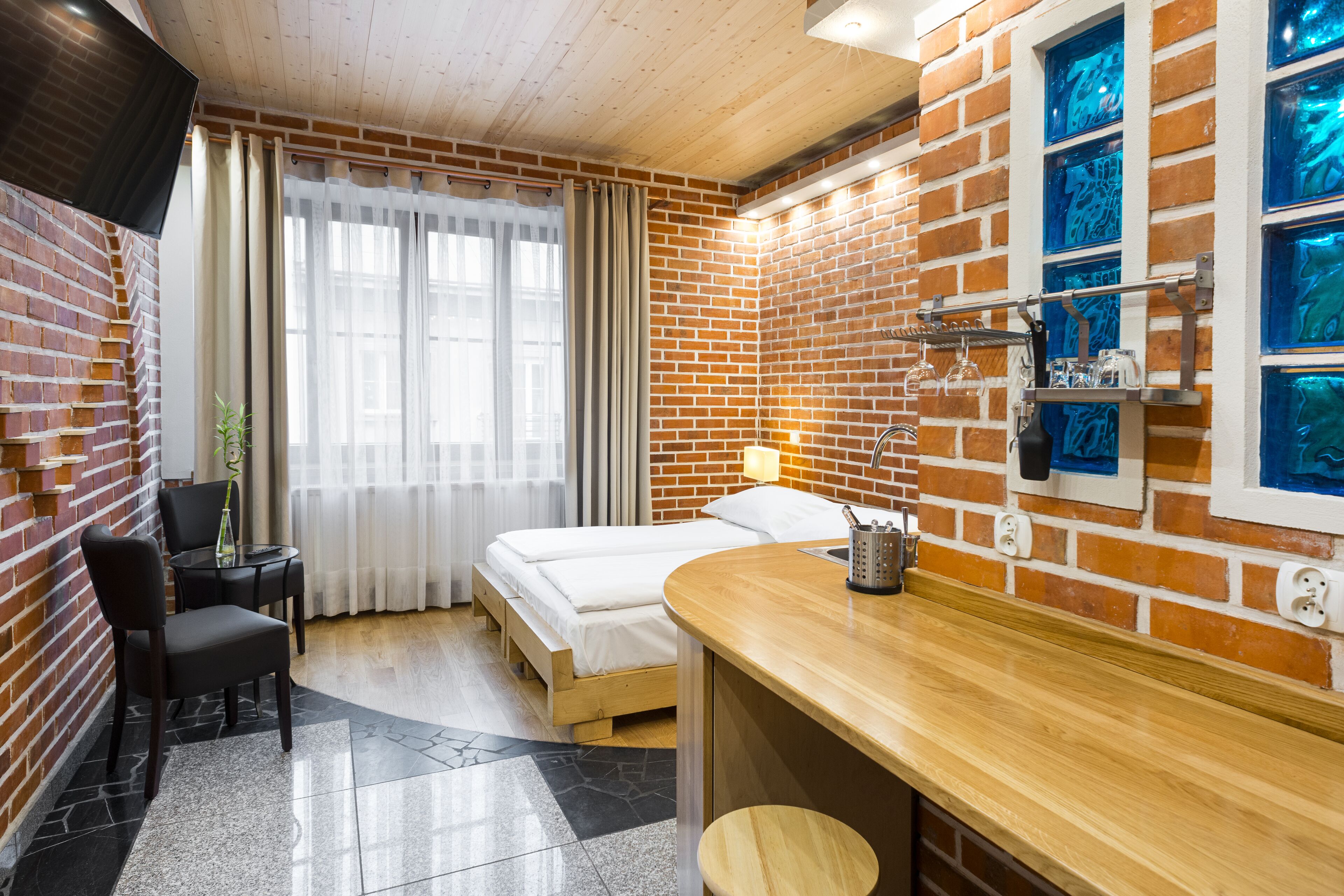 A hotel room with a brick wall, a wooden desk, a bed, and a window with blue-tinted glass.