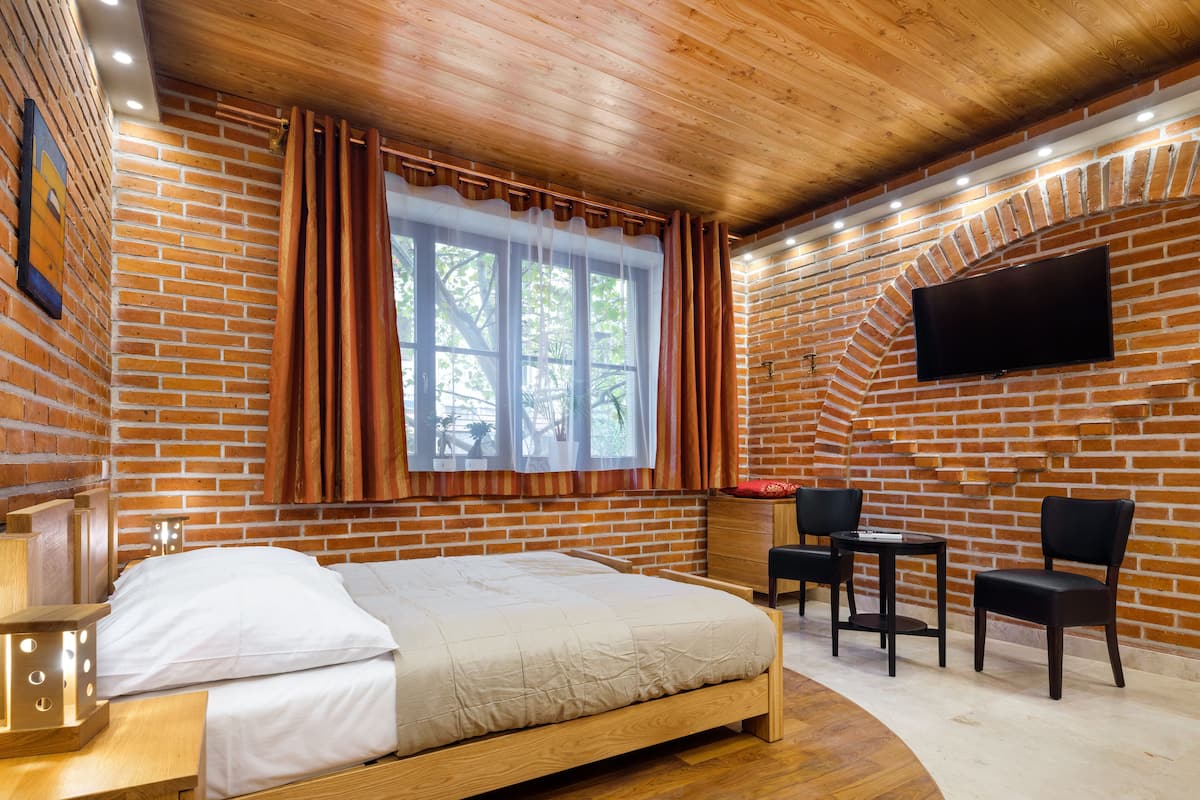 A room with a wooden bed, a desk with a chair, a TV mounted on the brick wall, and a window with curtains.