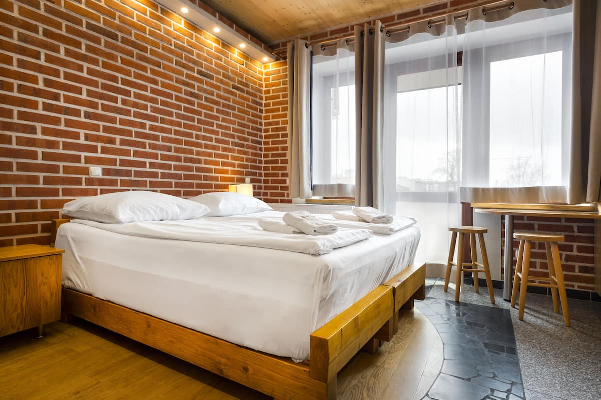 A room with a brick wall, a wooden bed, a desk, and a window with curtains.