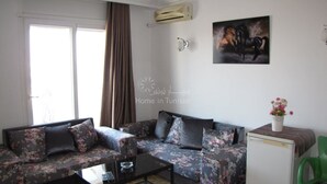 A living room with a sofa, a coffee table, and a small cabinet. There is a window with curtains, a wall-mounted air conditioner, and a painting of horses. - Superb apartment for vaccinations (sousse)