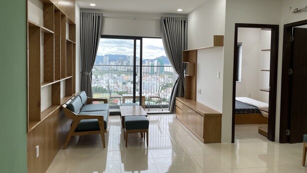 Una sala de estar moderna con un sofá, dos sillones, una mesa de centro y un televisor. Hay un ventanal con cortinas, vistas a un paisaje urbano y una estantería de madera.