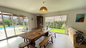 A spacious dining area with a wooden table and benches, large sliding glass doors leading to a backyard with a pool, and a sofa with a leopard print throw.