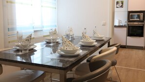 Un espace repas avec une longue table dressée pour un repas, des chaises, un coin cuisine équipé d’appareils électroménagers et une fenêtre avec des stores.