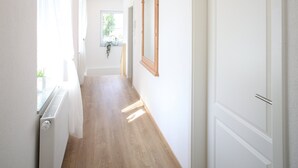 Un couloir avec un plancher en bois, des murs blancs, un radiateur, une porte et une fenêtre avec des rideaux.