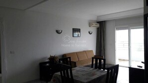 A living room with a sofa, a dining table with chairs, and a large window with curtains. - Great waterfront apartment (Sousse)