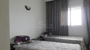 A room with two beds, a window with curtains, a wall-mounted light, and a small table. - Great waterfront apartment (Sousse)