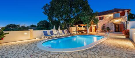 Une piscine avec des chaises longues, une terrasse en pierre et une maison en pierre avec un toit en tuiles.