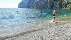 Ein Kiesstrand mit klarem Wasser, eine Person beim Paddleboarding und ein felsiger Klippenhintergrund.