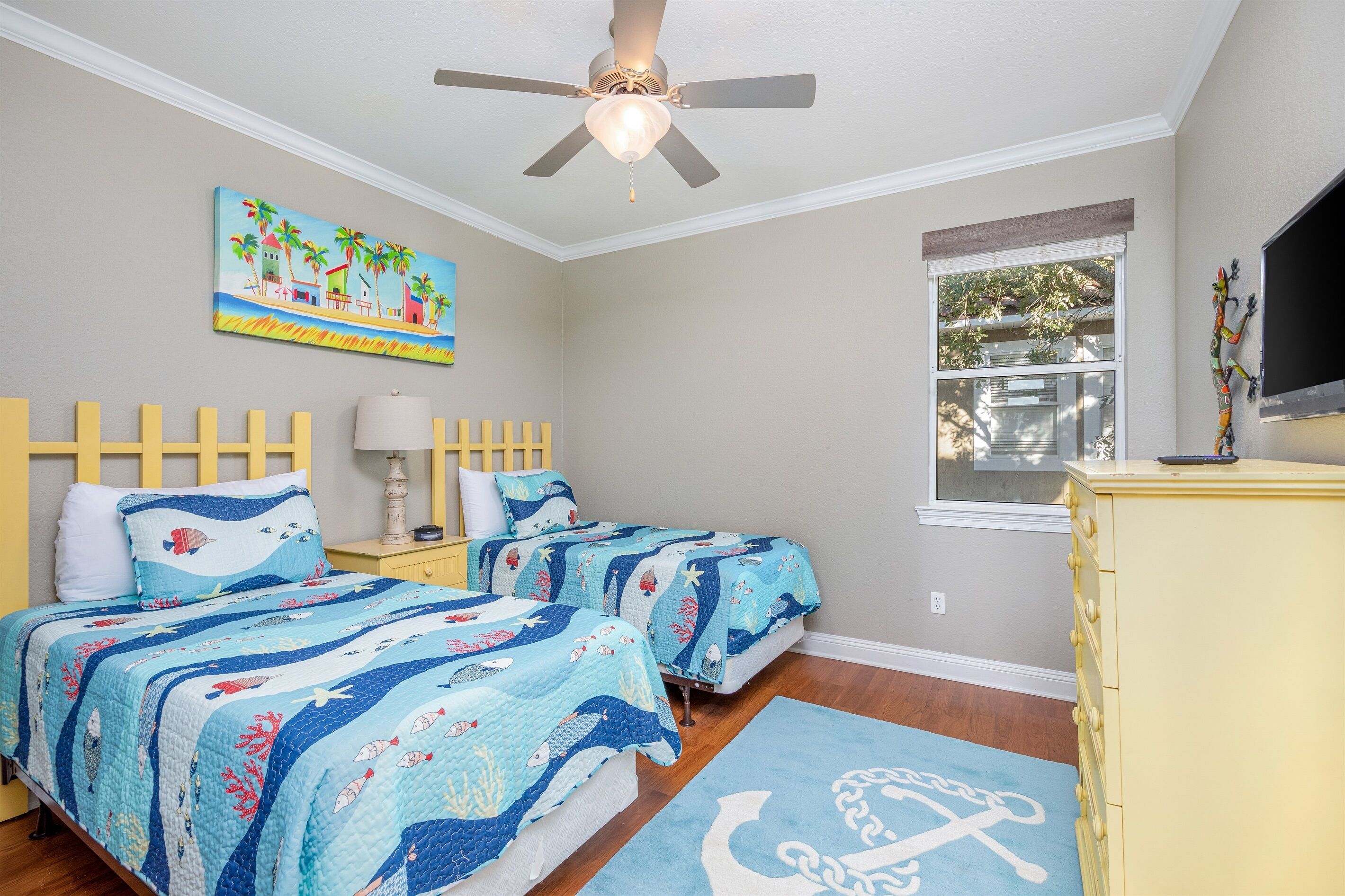 Un dormitorio con dos camas, ventilador de techo, pintura con temática de playa y ventana con vistas al exterior.