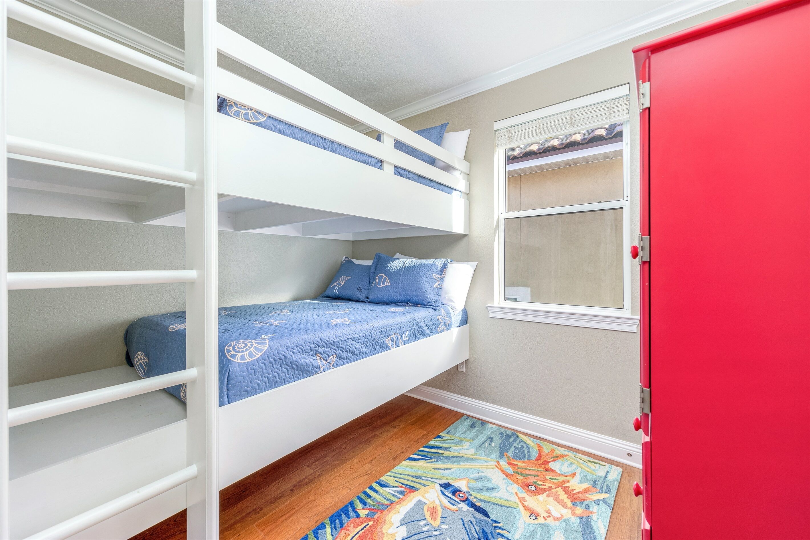 A bunk bed room with a red door, a window with blinds, and a colorful rug.