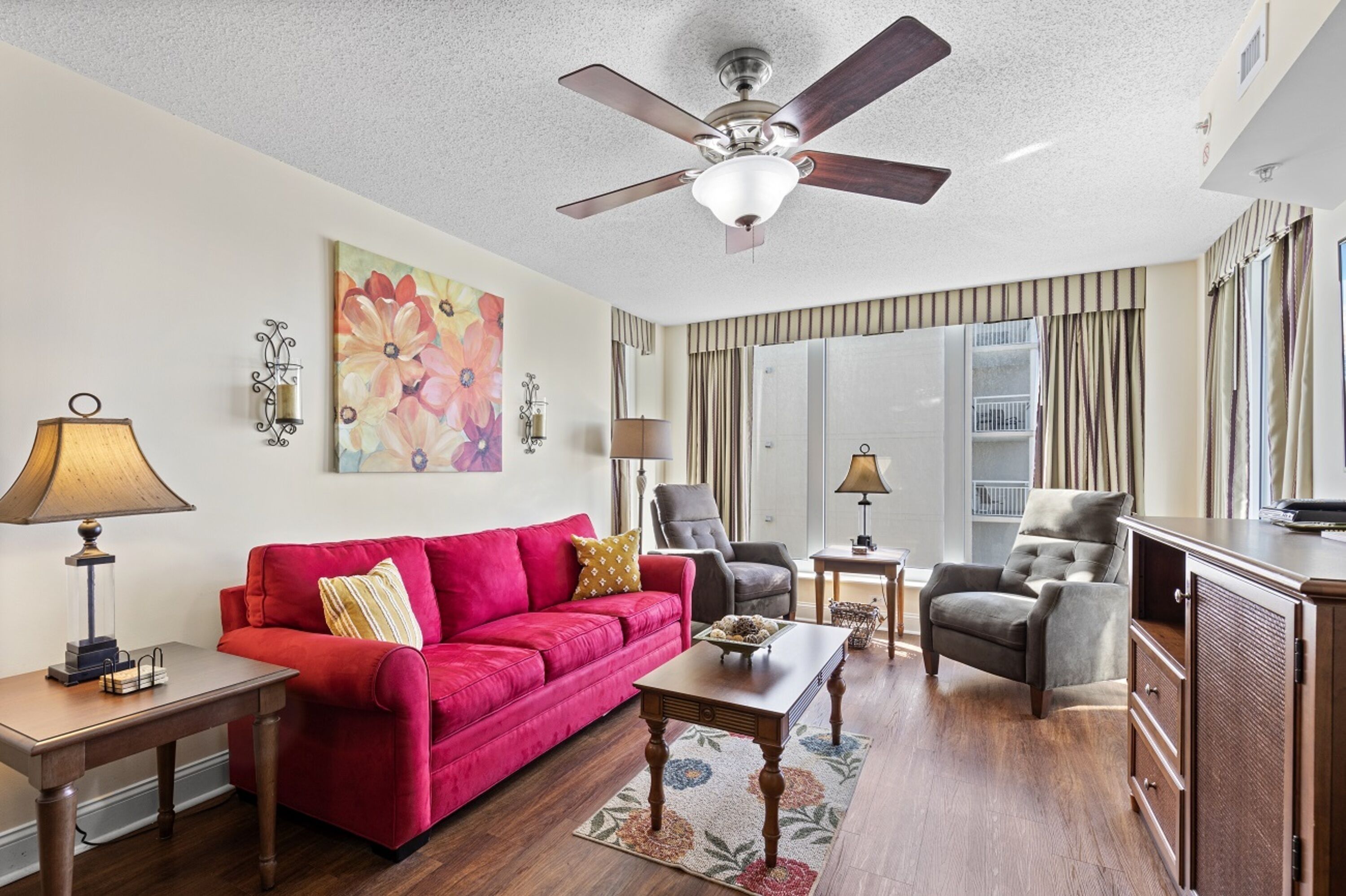 Une chambre d’hôtel avec un canapé rouge, une table basse en bois, un fauteuil gris, un ventilateur de plafond et une grande fenêtre avec des rideaux.