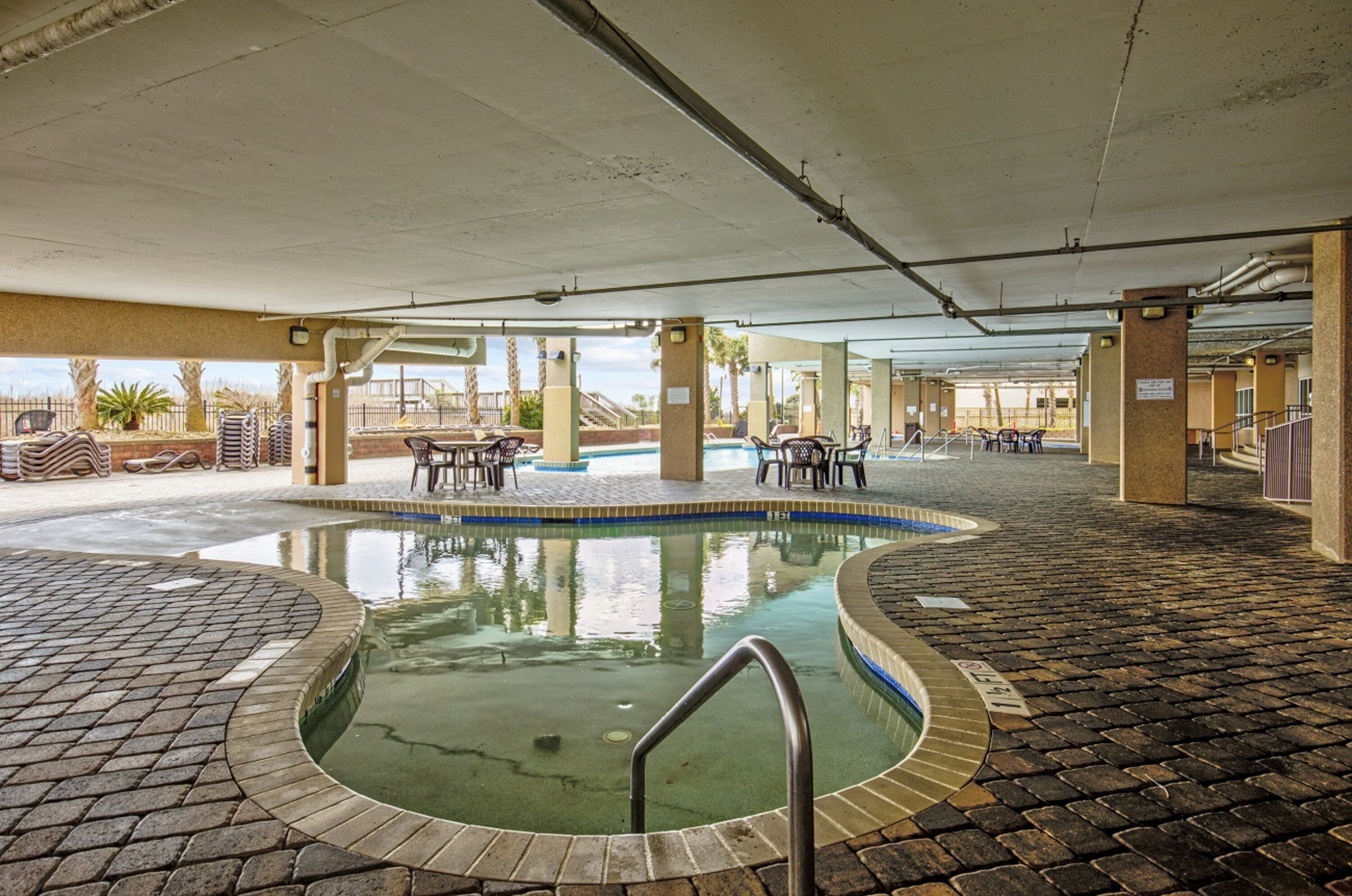 A covered outdoor swimming pool with a brick border, surrounded by lounge chairs and tables, and a view of palm trees and a building in the background.