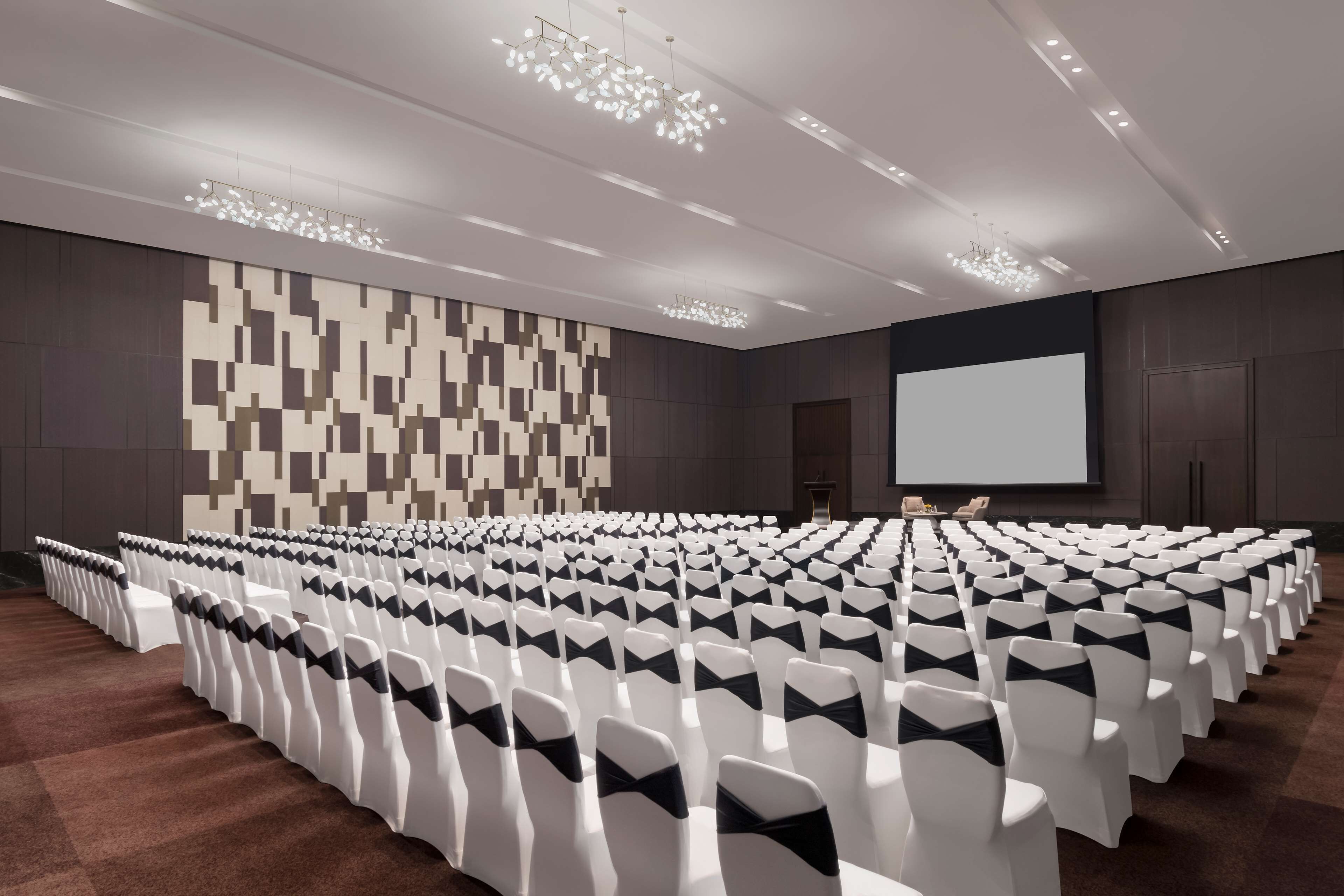 A conference room with rows of white chairs, a large screen, and a patterned wall.