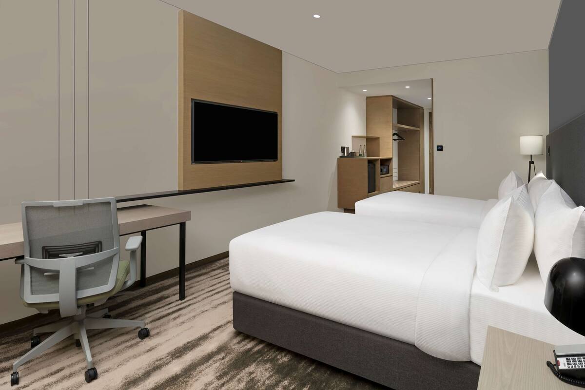 A modern hotel room with a large bed, a desk with a chair, a flat-screen TV mounted on the wall, and a bedside table with a lamp.