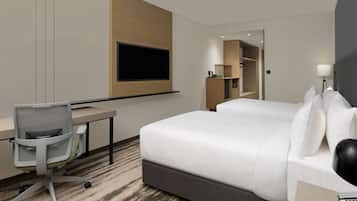 A modern hotel room with a large bed, a desk with a chair, a flat-screen TV mounted on the wall, and a bedside table with a lamp.