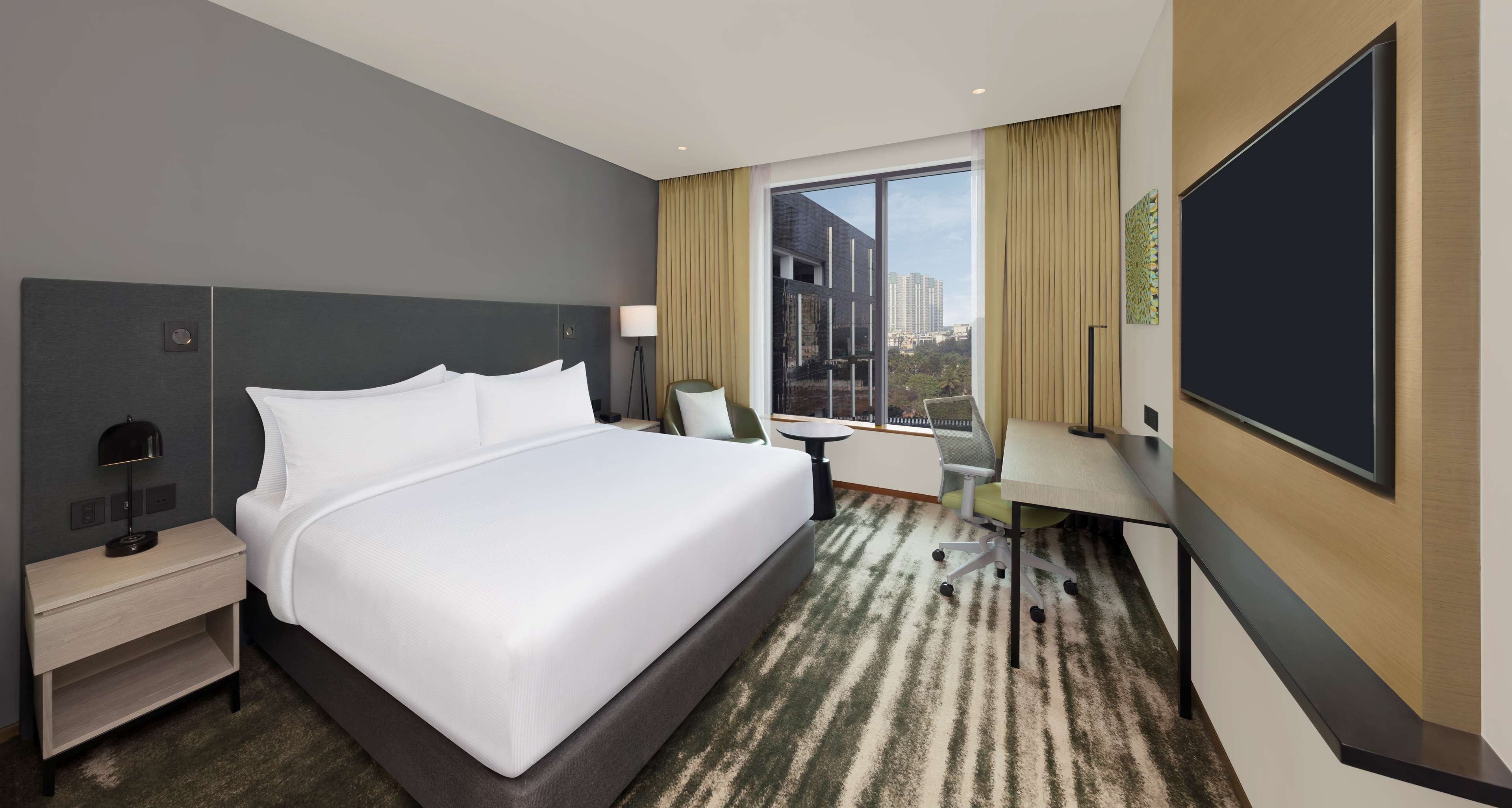 A hotel room with a large bed, a desk, a chair, a TV, and a window with a city view.