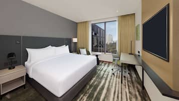 A hotel room with a large bed, a desk, a chair, a TV, and a window with a city view.