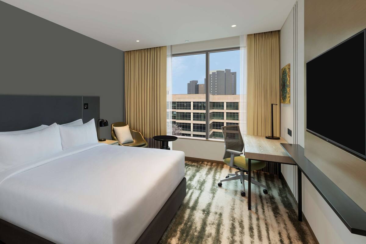A hotel room with a large bed, a desk, a chair, a TV, and a window with a city view.