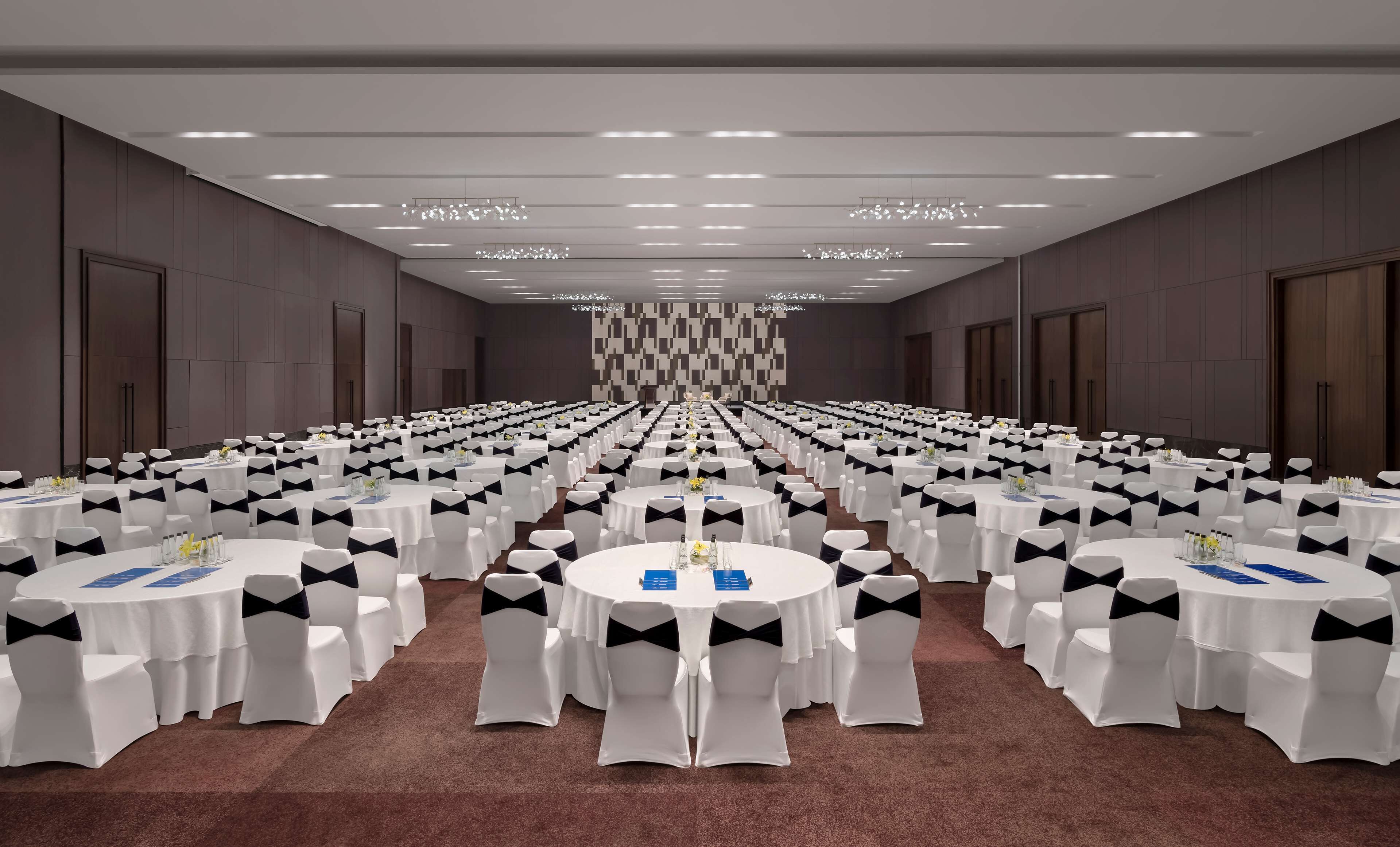 A large conference room with rows of round tables, each with white tablecloths and black chair covers, and a carpeted floor.