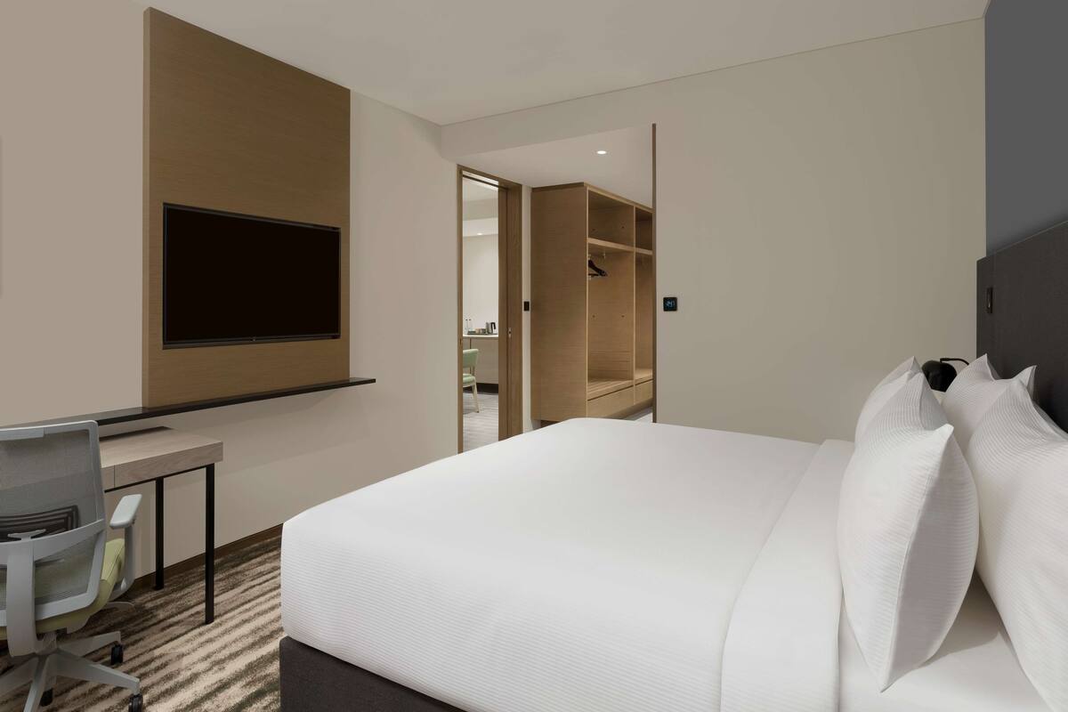 A hotel room with a bed, a desk with a chair, a TV mounted on the wall, and a wardrobe.