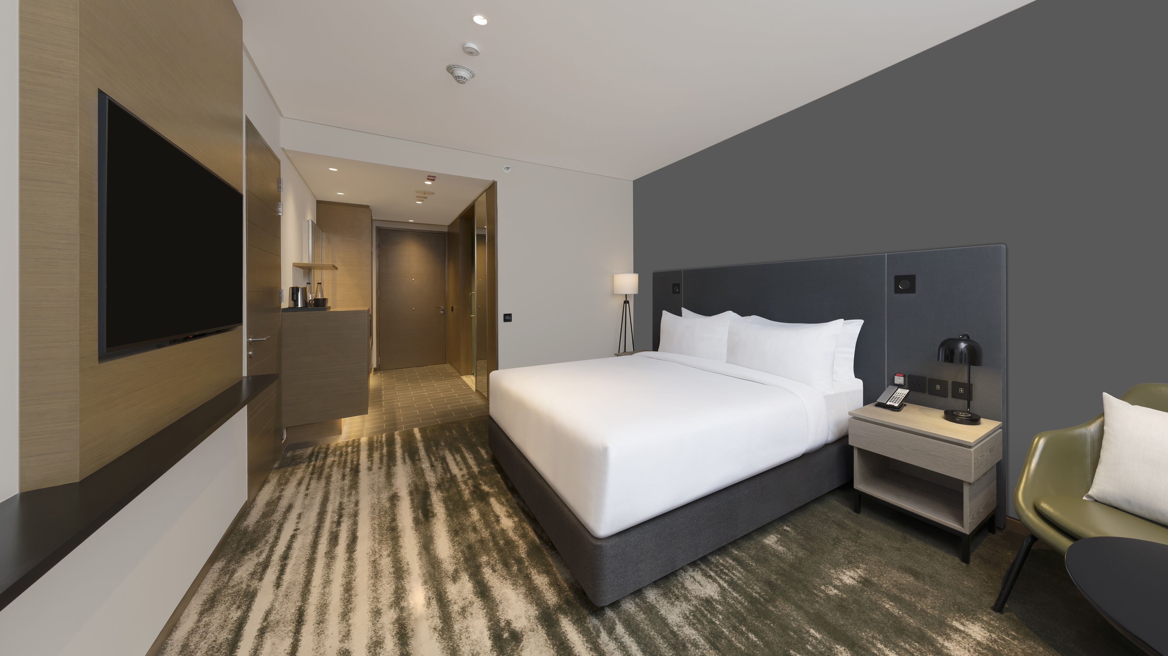 A modern hotel room with a large bed, bedside table, lamp, and a TV mounted on the wall.