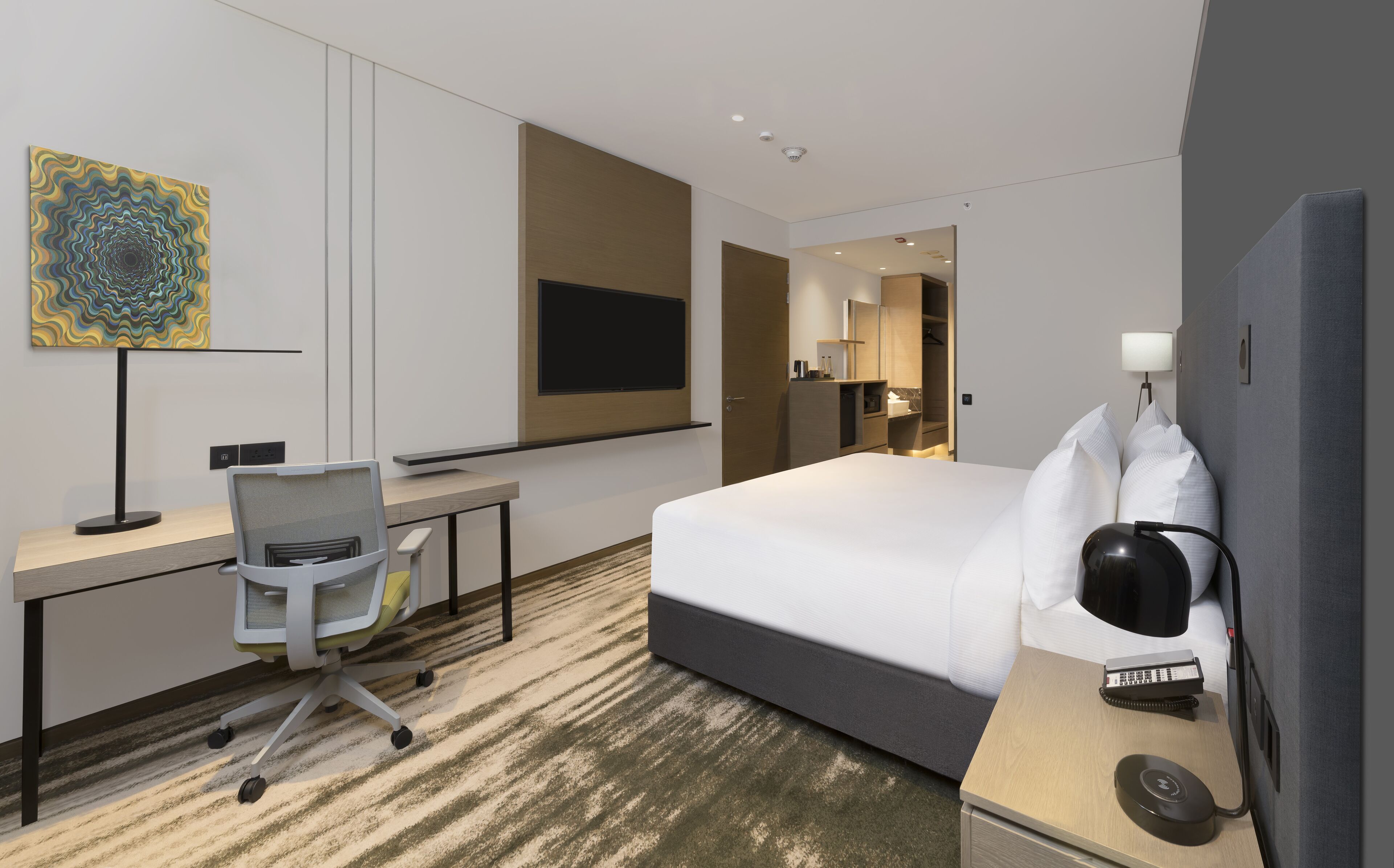 A hotel room with a bed, desk, chair, and a large artwork on the wall.
