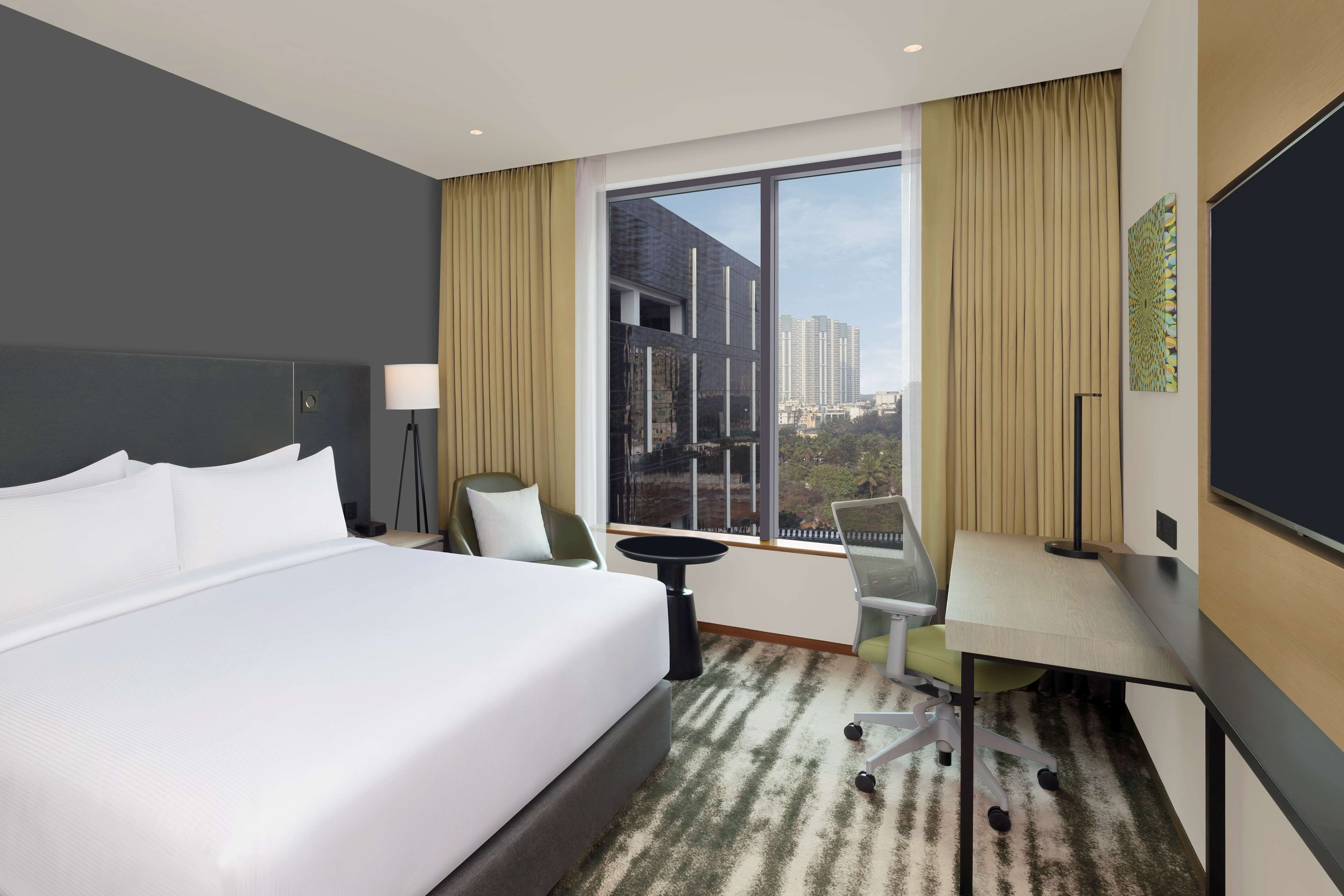 A modern hotel room with a large bed, a desk, a chair, and a view of the cityscape through large windows.