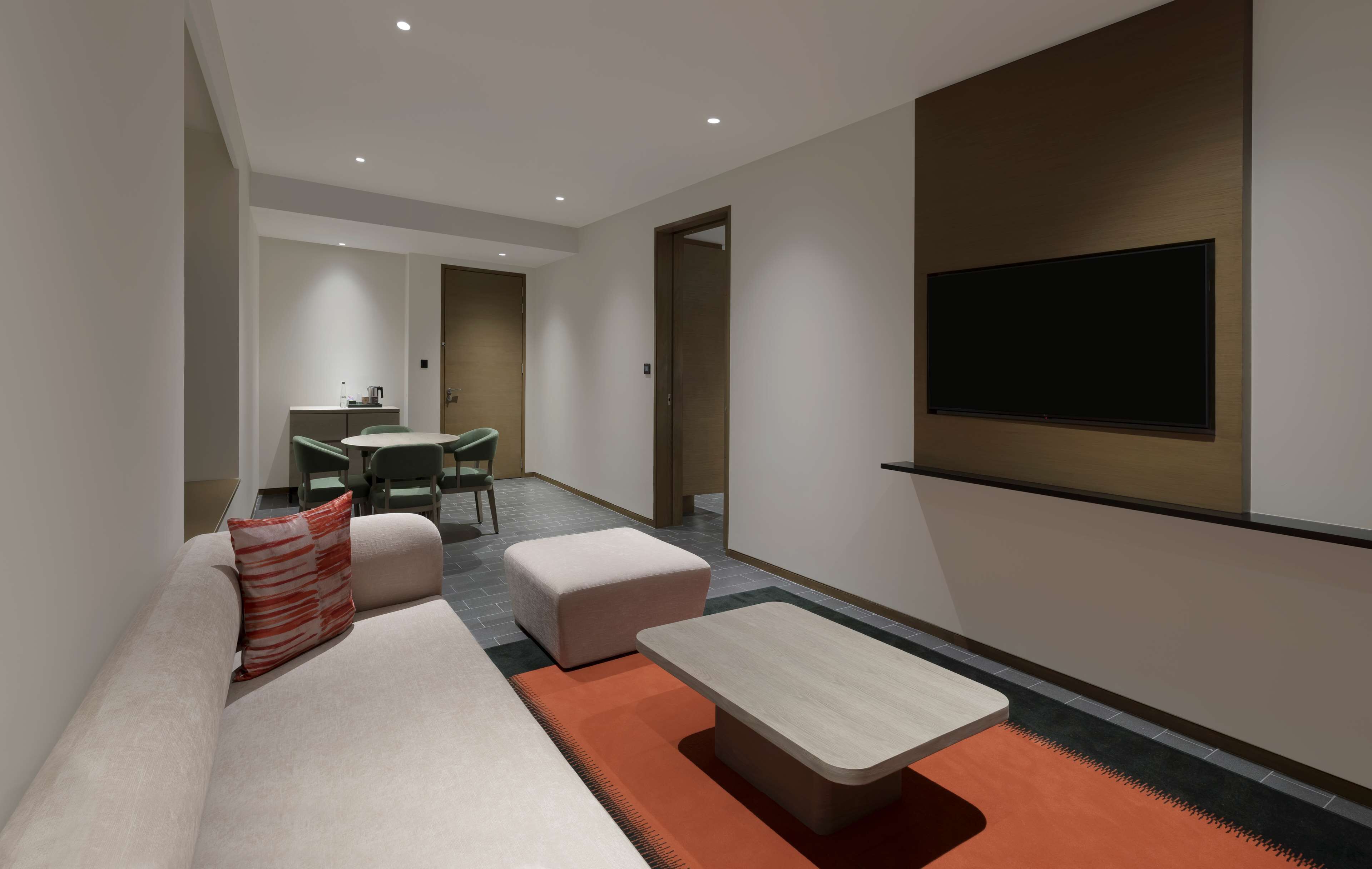 A modern living room with a sofa, ottoman, and coffee table. There is a dining area with a table and chairs, and a kitchen in the background.