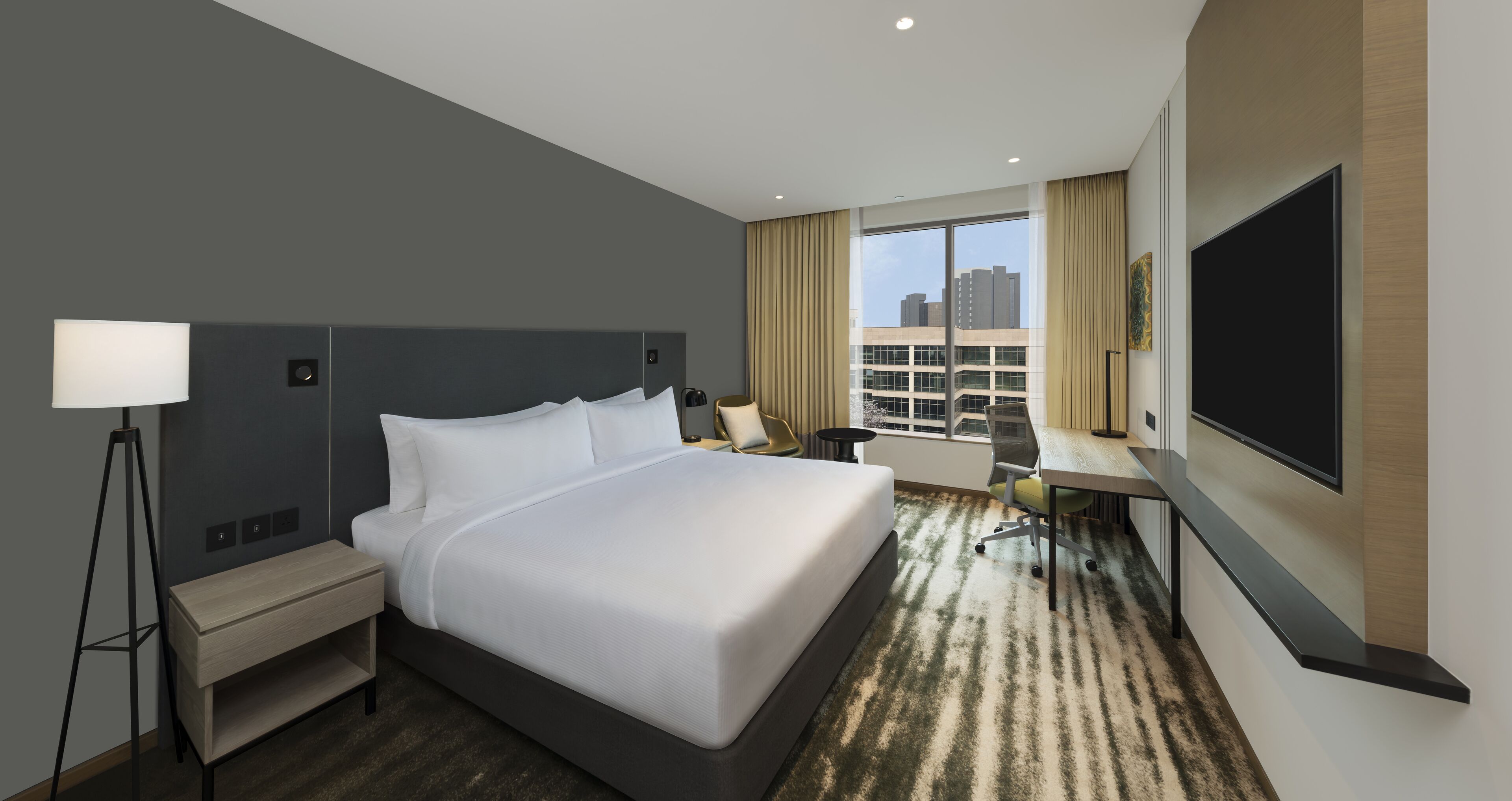 A hotel room with a large bed, a desk, a chair, a TV, and a window with a city view.