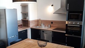 Une cuisine moderne avec une table en bois, des chaises noires, un luminaire suspendu et des appareils électroménagers intégrés, notamment un lave-vaisselle et un four.