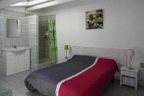 Un dormitorio con cama, mesita de noche, espejo y baño con lavamanos y ducha.