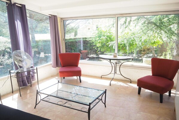 A room with a glass table, two red chairs, a fan, and a view of a garden through large windows.
