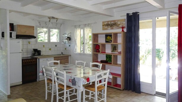 Una cocina con una mesa de comedor y sillas, un refrigerador, estufa y microondas. Hay una ventana con cortinas, un estante con objetos decorativos y una puerta que da a un área exterior.