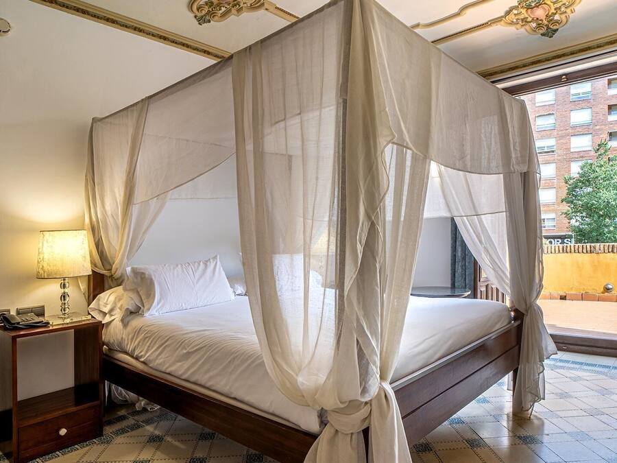 A four-poster bed with sheer curtains, a bedside lamp, and a view of a building through a window.