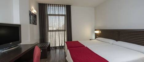 A hotel room with a bed, a desk with a TV, a chair, and a window with curtains.
