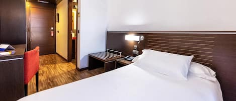 A hotel room with a bed, bedside table, chair, and a desk with a chair.