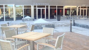 Een terras met tafels en stoelen, een overdekt zitgedeelte, en een gebouw met de borden 'CAFFETERIA' en 'RESTAURANT'.