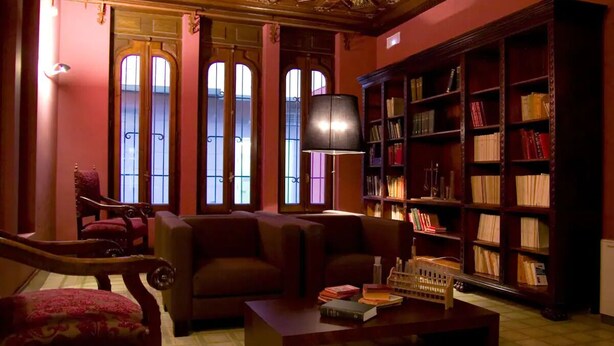 Een knusse bibliotheek met een grote boekenkast, twee banken en een tafel met boeken en een modelhuis.