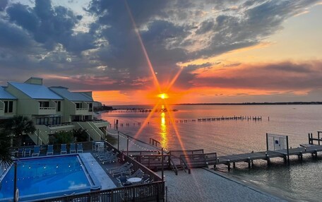 A sunset over a calm body of water with a dock extending into the water. There is a pool area with lounge chairs and a building with a sloped roof in the foreground.